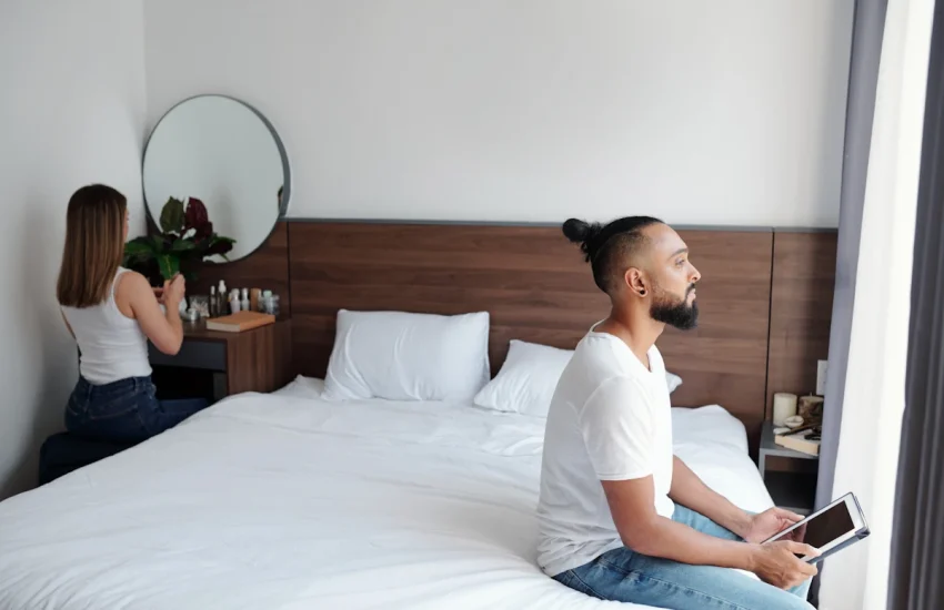 Un homme assis d'un côté d'un lit et une femme qui se regarde dans un miroir à côté